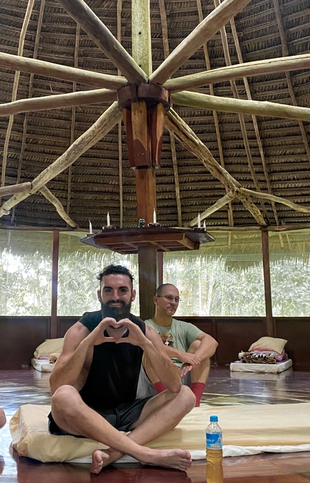 Happy male Caya Shobo guest makes heart sign with hands sitting in the traditional Shipibo maloka before Ayahuasca Ceremony